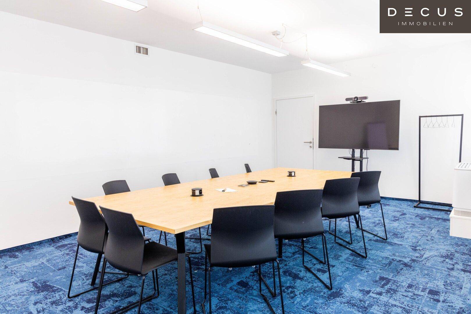 Großer Konferenzraum mit einem langen Holztisch, schwarzen Stühlen und einem großen Wandbildschirm für Präsentationen.