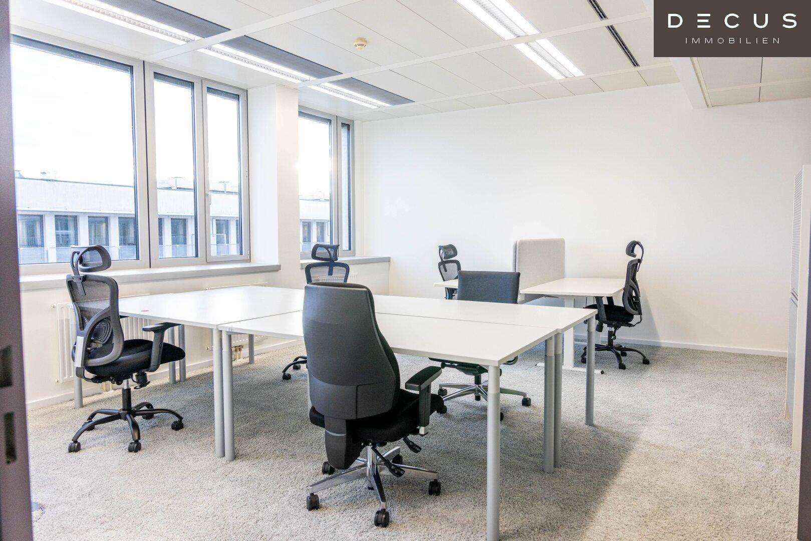 Geräumiger Bürobereich mit weißen Schreibtischen, ergonomischen Stühlen und großen Fenstern, die viel Licht hereinlassen.