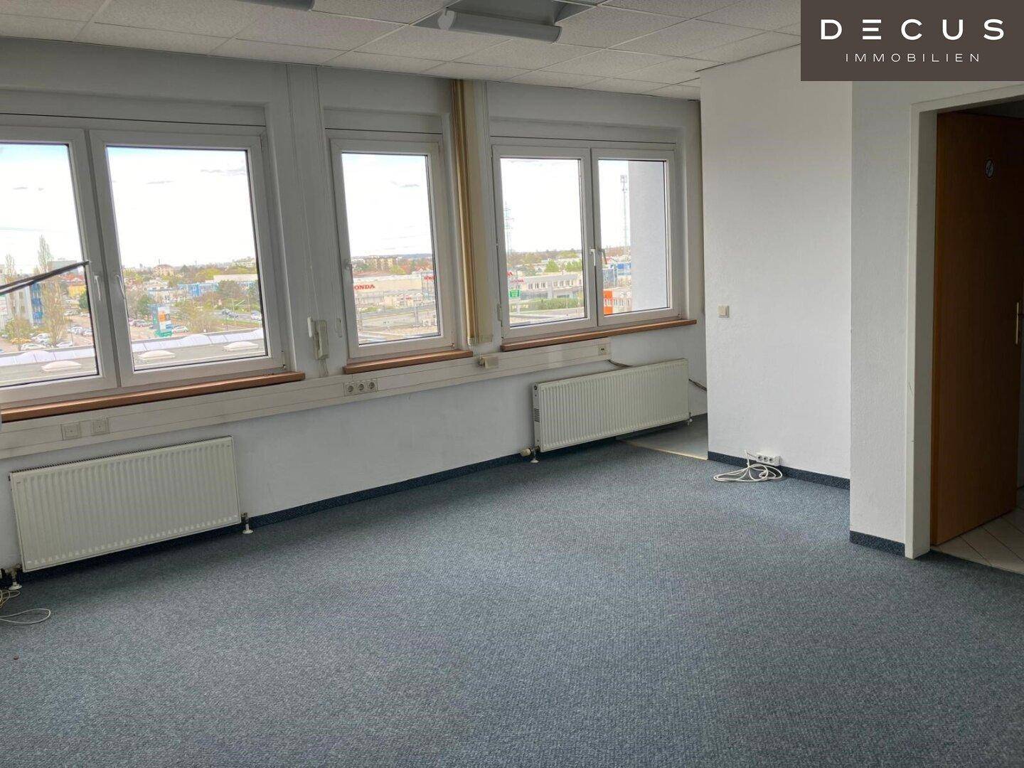 Leeres Büro mit grauem Teppichboden, großen Fenstern und Heizkörpern, bietet einen Blick auf die Stadt.