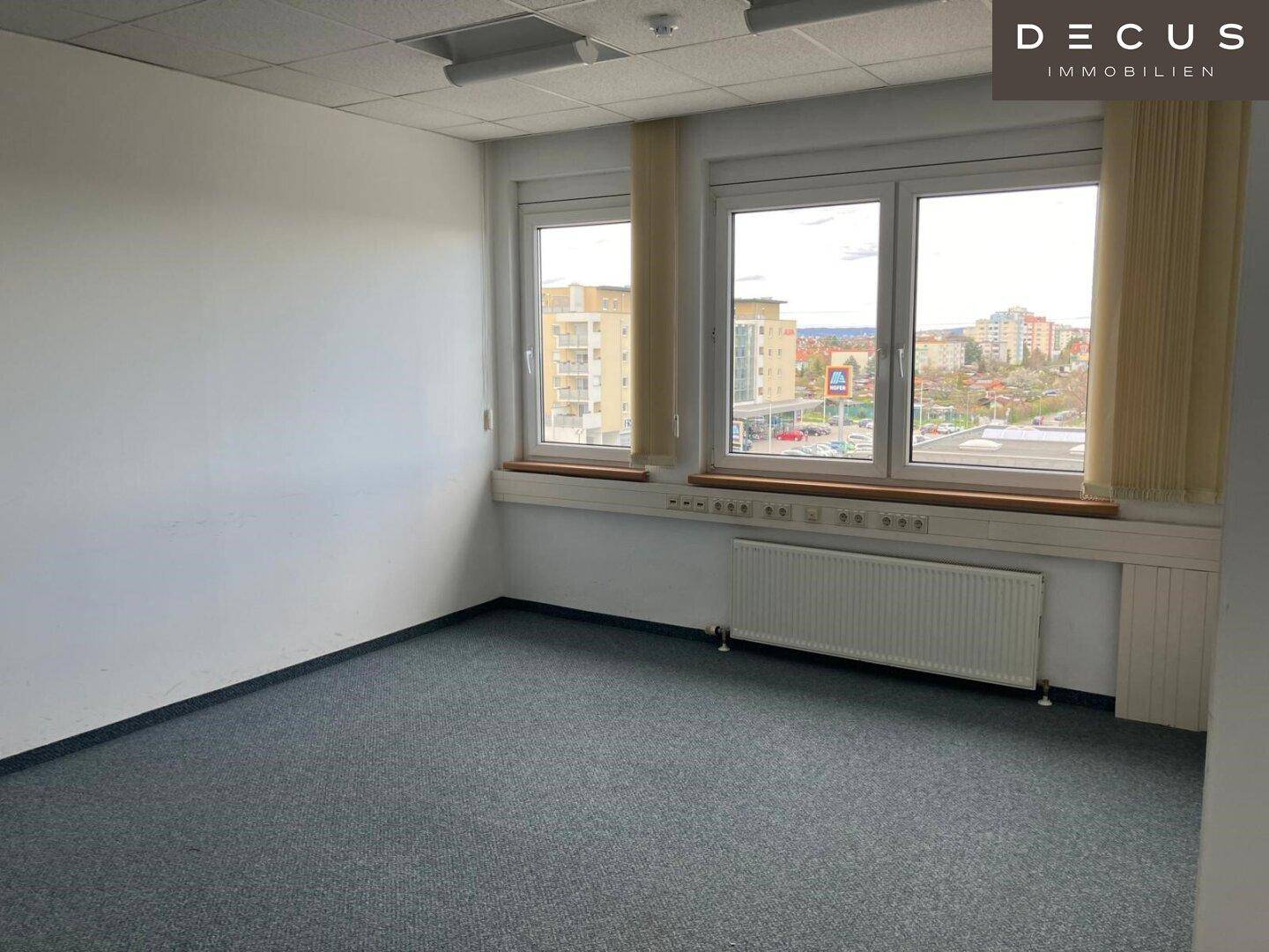 Helles Büro mit Teppichboden und mehreren Fenstern, die viel Tageslicht hereinlassen und eine städtische Aussicht bieten.