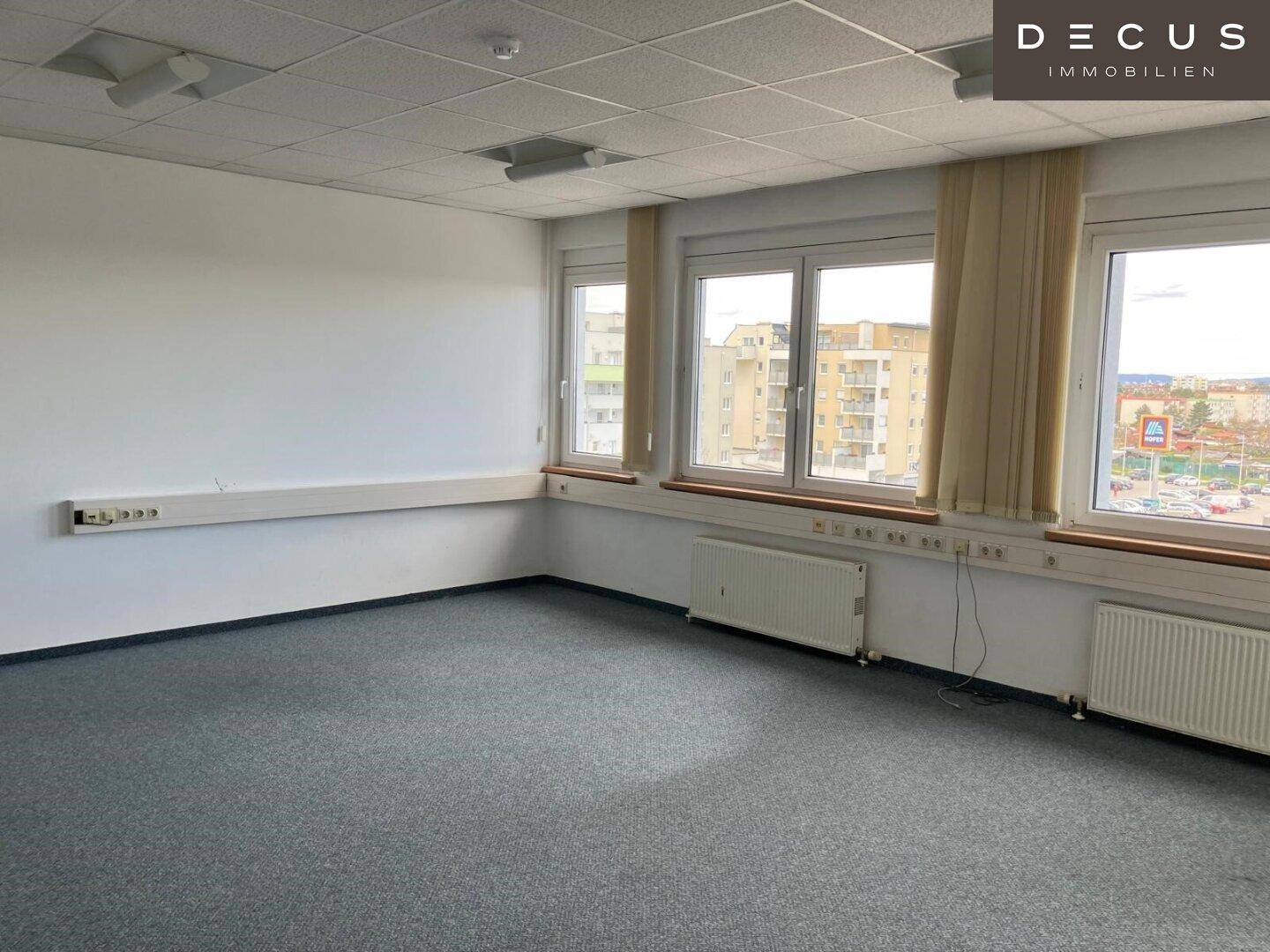 Geräumiges Büro mit grauem Teppichboden, großen Fenstern und Heizkörpern, bereit zur individuellen Gestaltung.