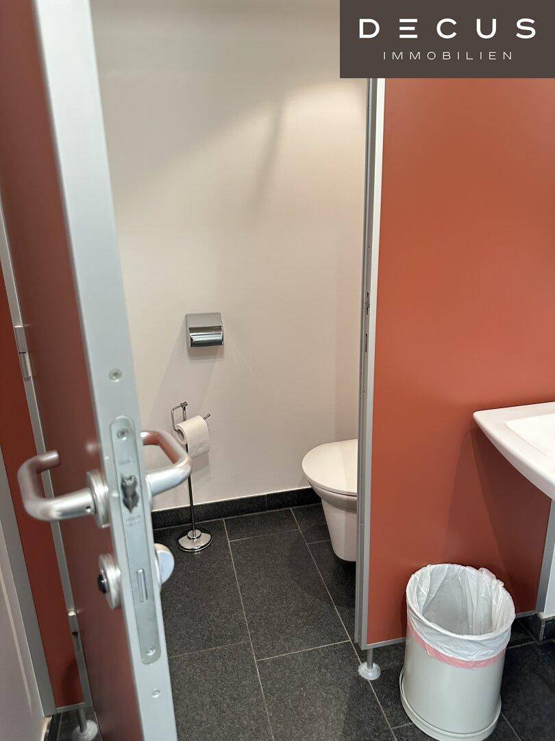 Kompakte Toilette mit modernen Trennwänden, Wand-WC und dunklen Fliesen, gut beleuchtet.
