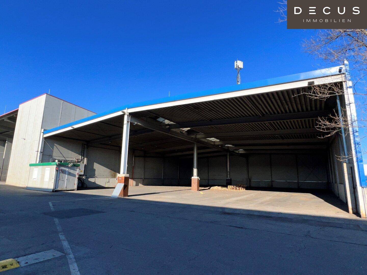 Weitläufige Industriehalle mit offener Bauweise und blauer Dachkante, umgeben von einem großen Vorplatz.