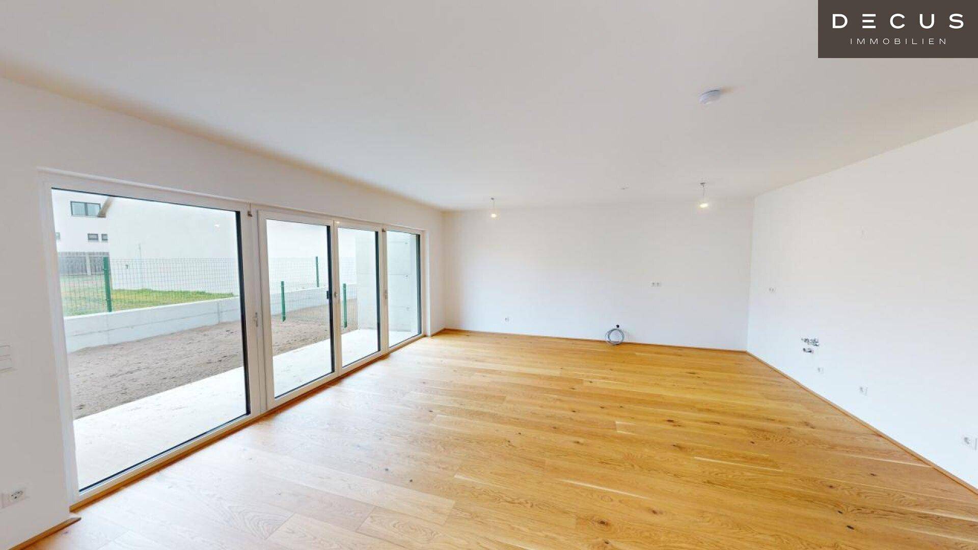 Geräumiges Wohnzimmer mit Holzboden und breiter Fensterfront, die viel Tageslicht hereinlässt.