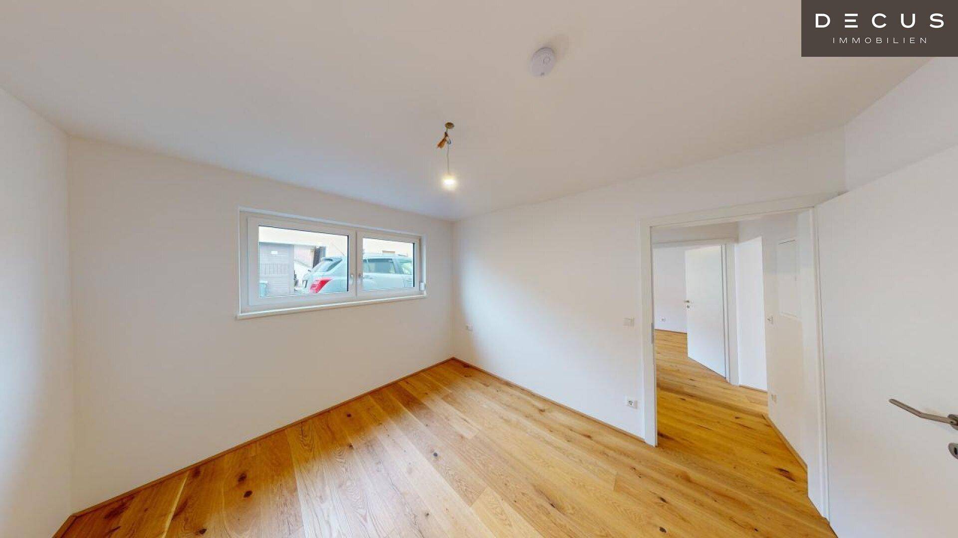 Leeres Zimmer mit Holzboden und einem Fenster, das den Blick nach draußen freigibt.