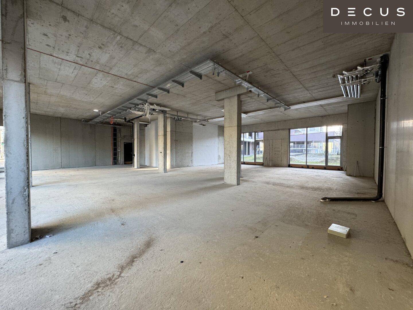 Geräumiger Gewerberaum im Rohzustand mit freiliegenden Installationen und Betonoberflächen.