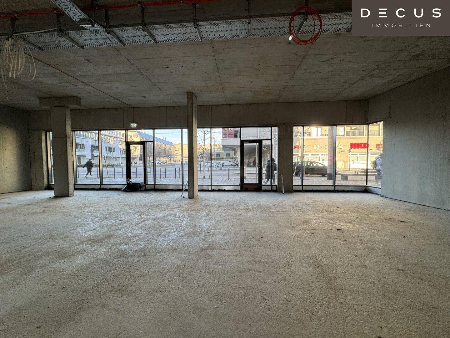 Großer, offener Gewerberaum im Rohbau mit Betonboden und raumhohen Fenstern zur Straße.