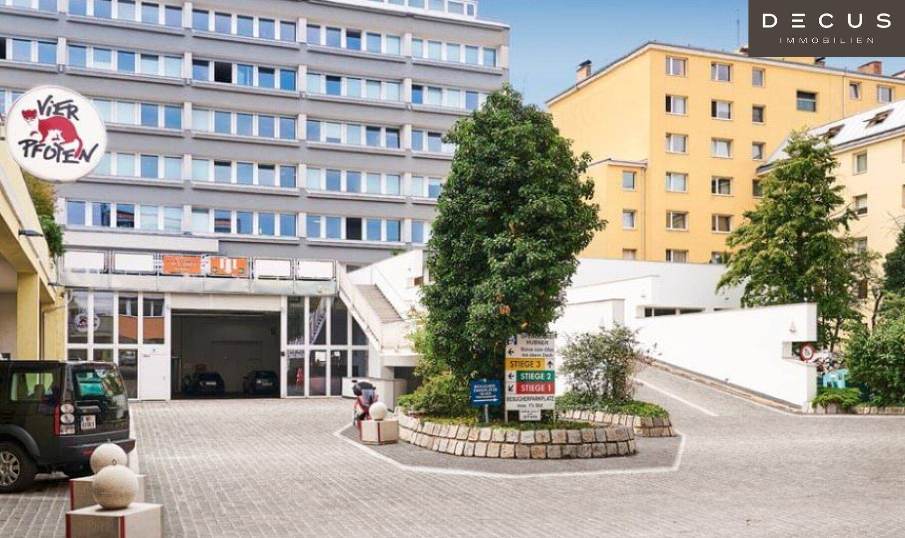 Zufahrt zu einem Parkhaus mit einem Baum in der Mitte und umliegenden Gebäuden.