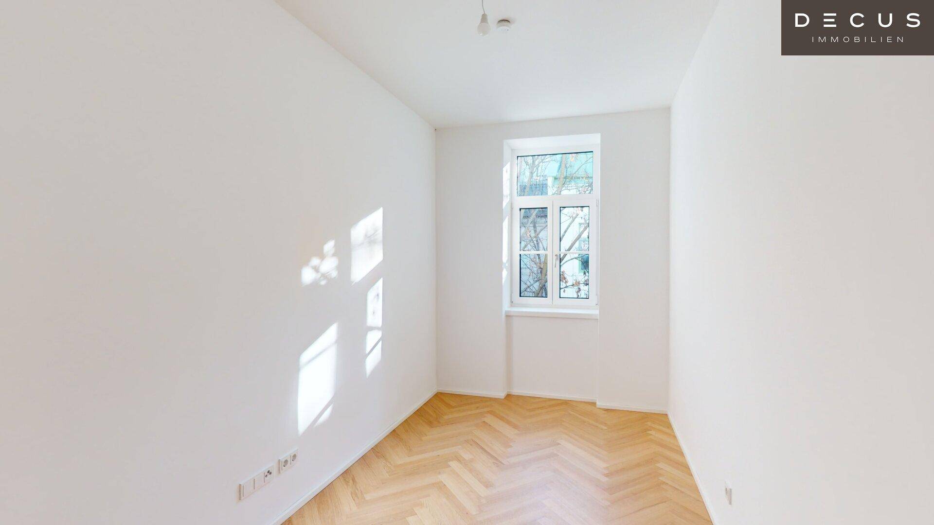 Helles Zimmer mit Fischgrätparkett und einem Fenster, das viel Tageslicht hereinlässt.