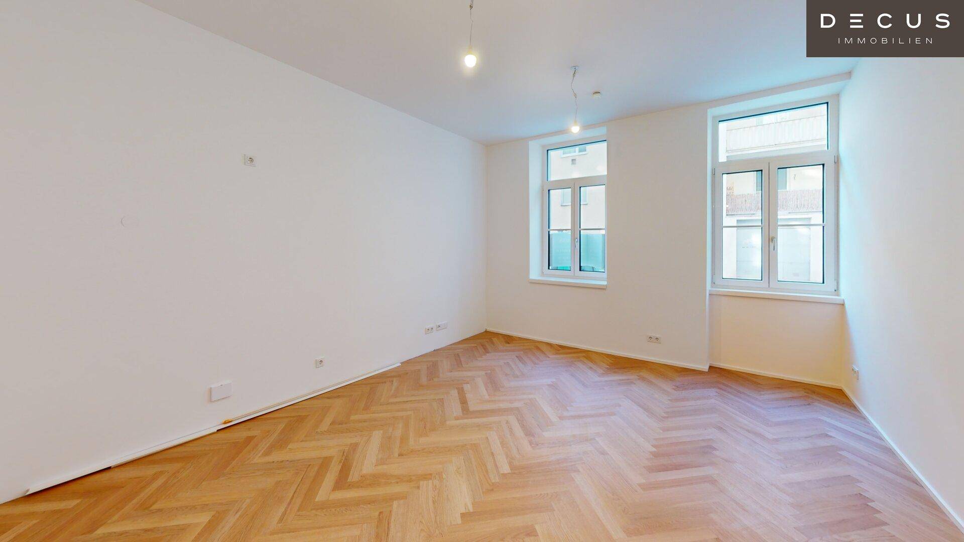 Geräumiges Zimmer mit Fischgrätparkett, hohen Decken und zwei großen Fenstern.