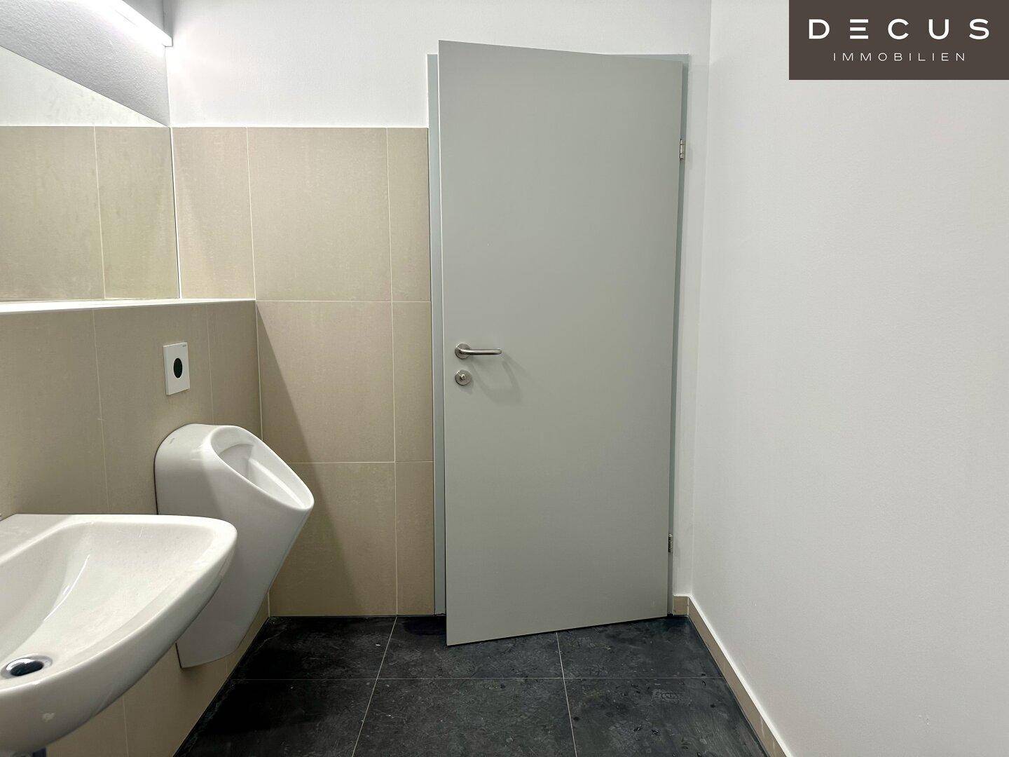 Modernes Herren-WC mit Urinal, Waschbecken und dunklen Fliesen auf dem Boden.