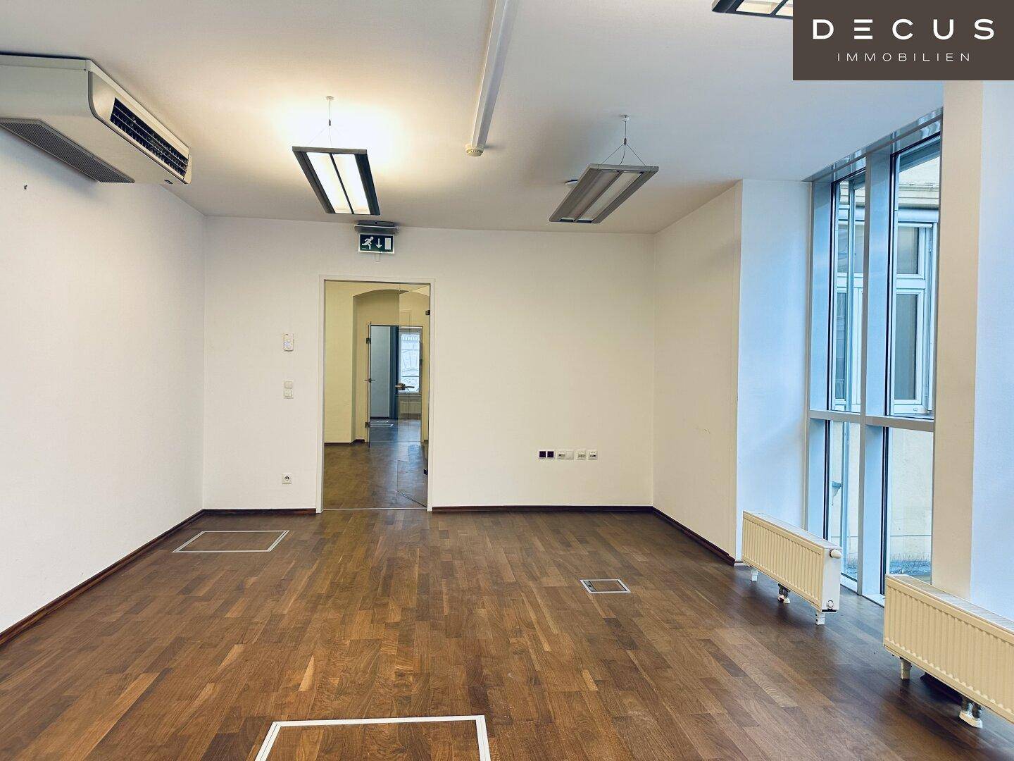 Leerer Büroraum mit Holzboden, großen Fenstern und Zugang zu weiteren Räumen, bietet vielseitige Nutzungsmöglichkeiten.