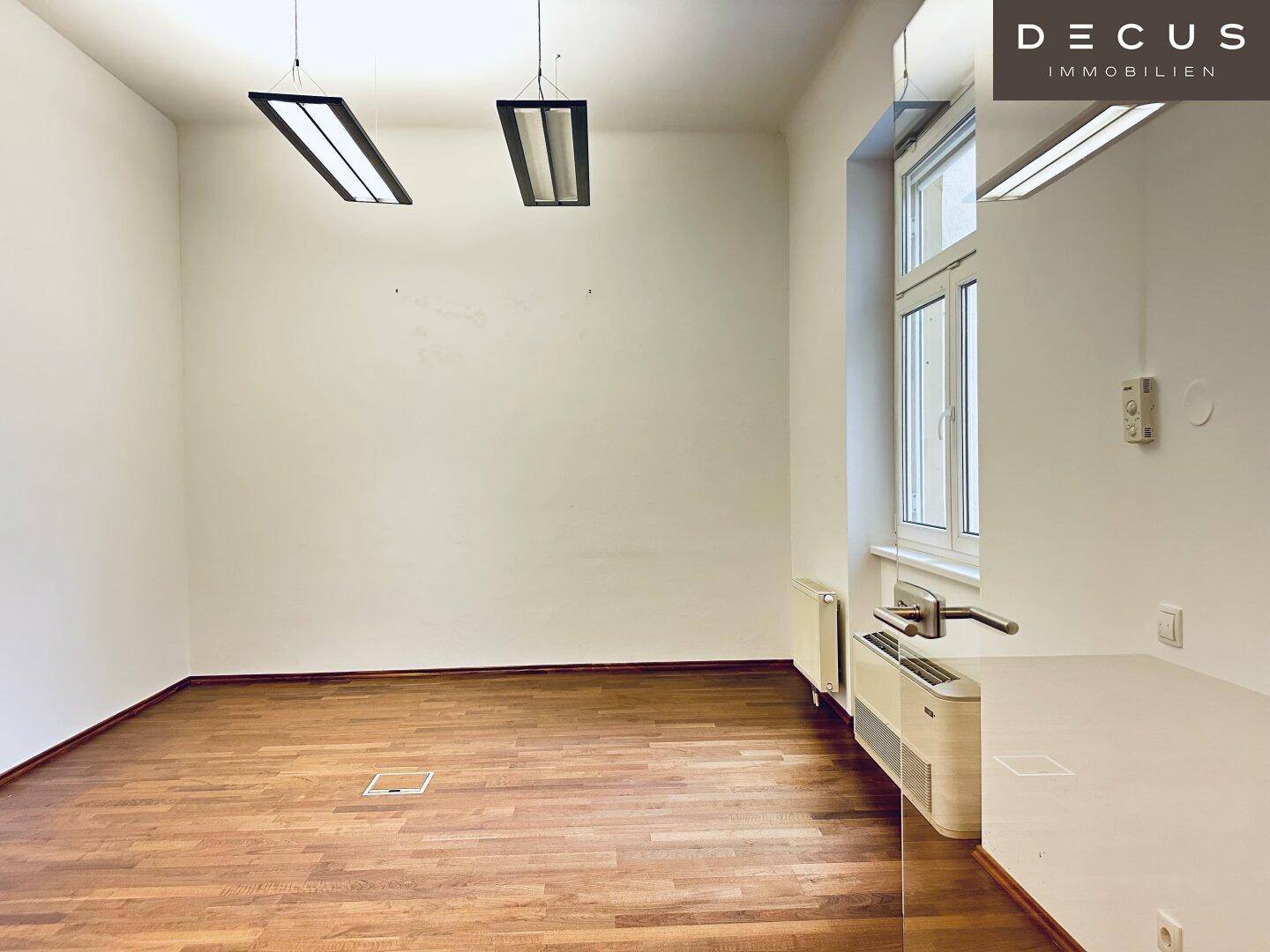 Ruhiger Büroraum mit Holzboden, großem Fenster und Deckenleuchten, ideal für konzentriertes Arbeiten.