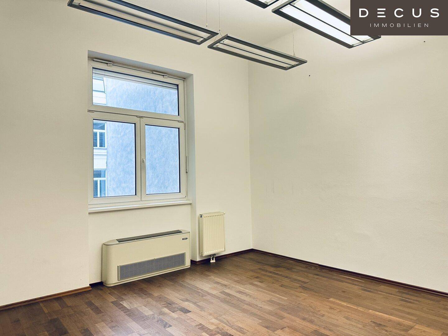 Heller Büroraum mit Holzboden und einem großen Fenster, das viel Tageslicht hereinlässt und eine angenehme Arbeitsatmosphäre schafft.