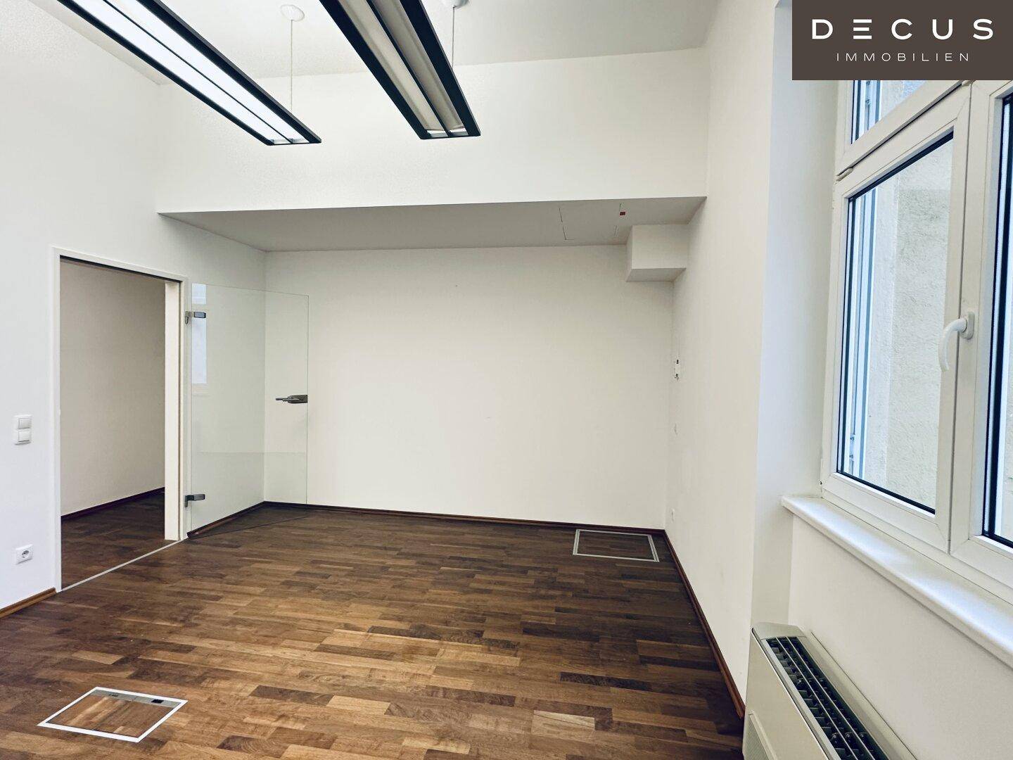 Geräumiger Büroraum mit Holzboden und einer modernen Glastür, die zu einem weiteren Bereich führt.