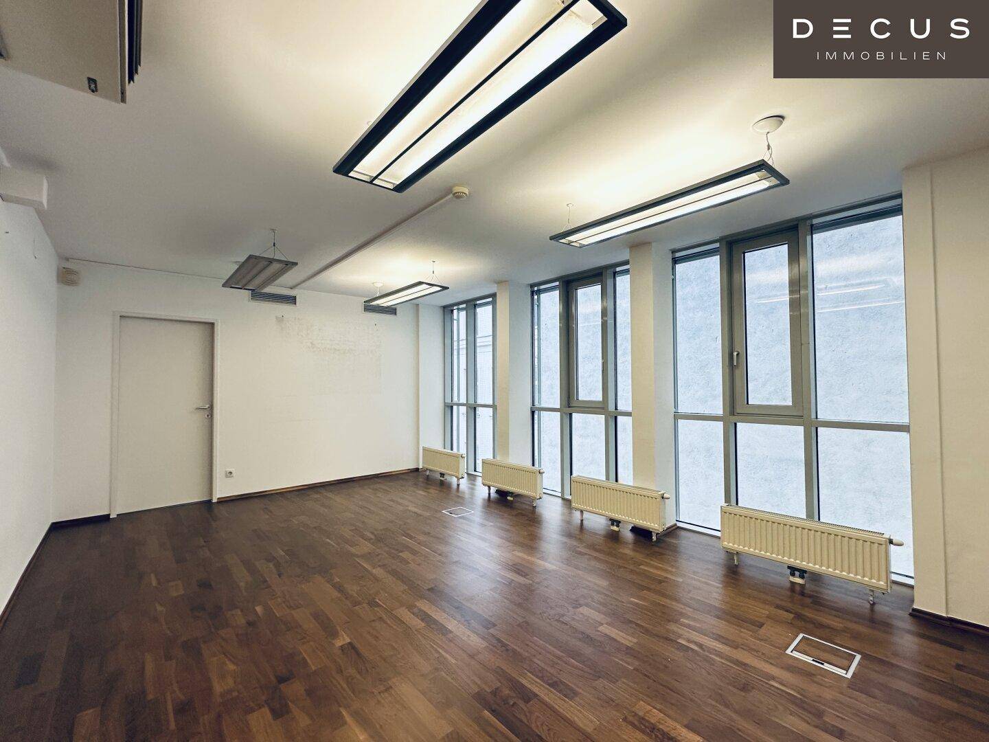 Großer Büroraum mit vielen Fenstern, Holzboden und Deckenleuchten, bietet eine helle und offene Arbeitsumgebung.