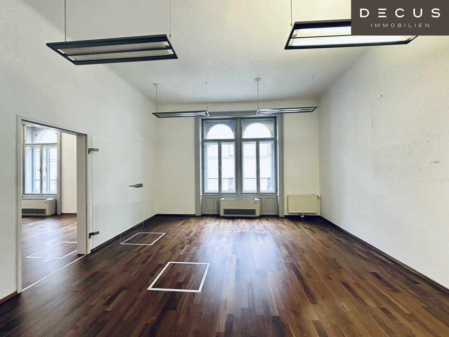 Heller Büroraum mit Holzboden, großen Bogenfenstern und einer modernen Glastür, die zu einem weiteren Bereich führt.