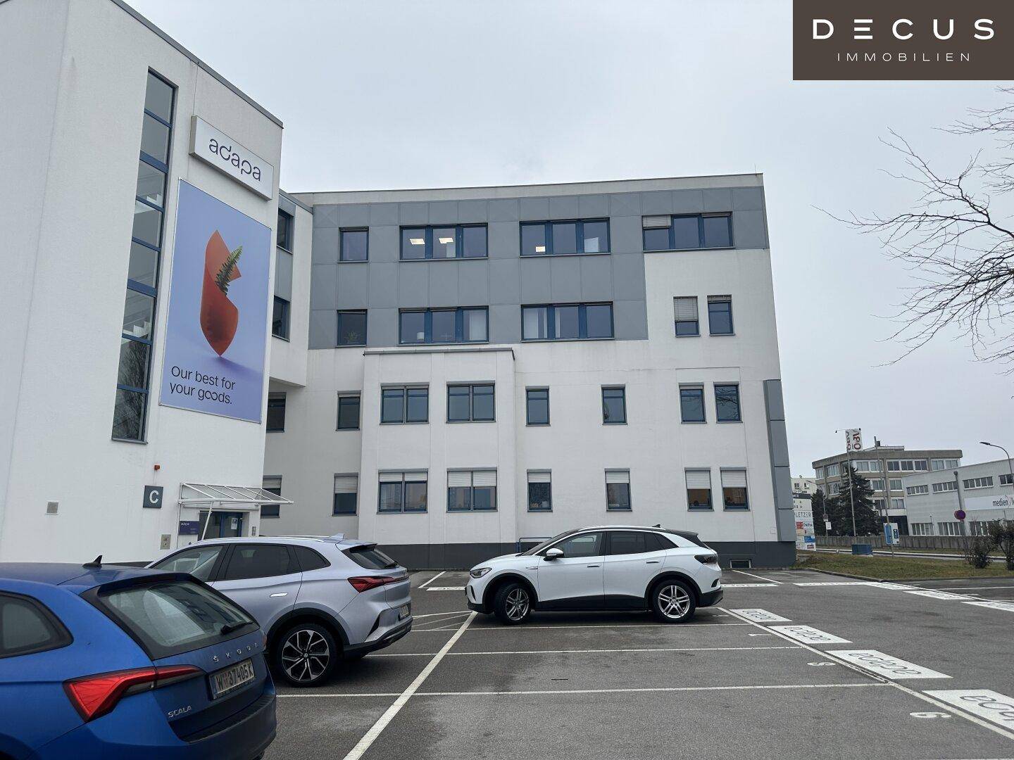 Moderne Bürogebäude-Fassade mit großen Fenstern und einem davorliegenden Parkplatz für Fahrzeuge.