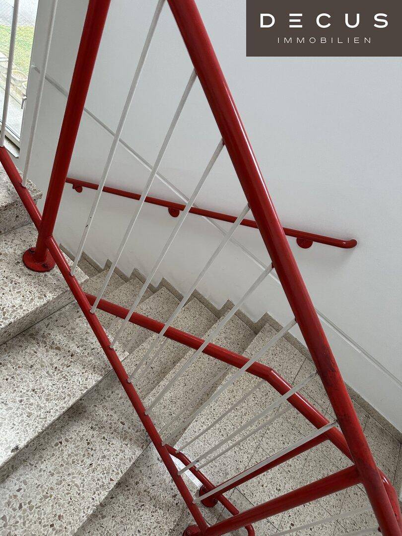 Treppenhaus mit Terrazzostufen und einem markanten roten Geländer, das zu den oberen Etagen führt.
