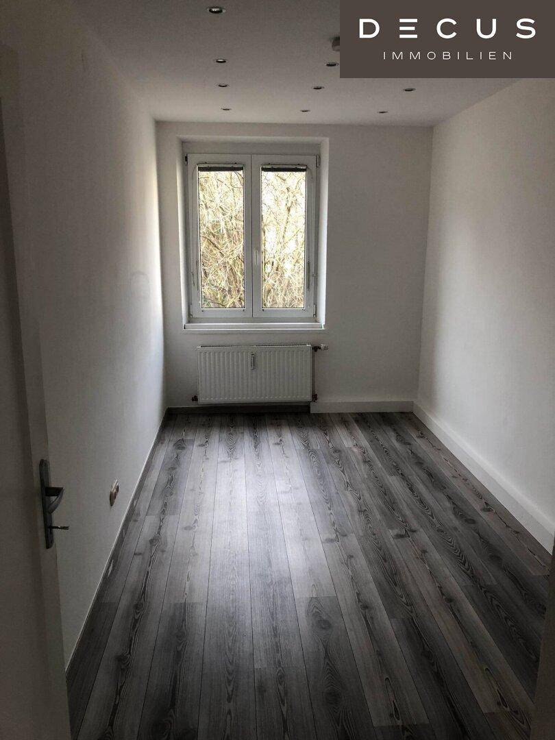 Kleines, helles Zimmer mit dunklem Laminatboden und Fenster mit Blick ins Grüne.