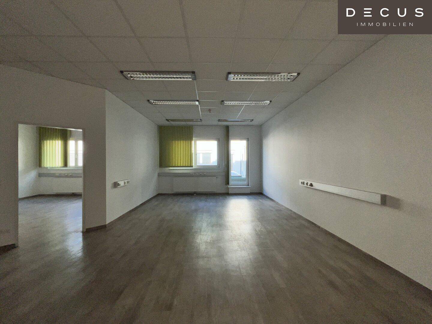 Heller Büroraum mit grauem Laminatboden und mehreren Fenstern, die viel Tageslicht hereinlassen.