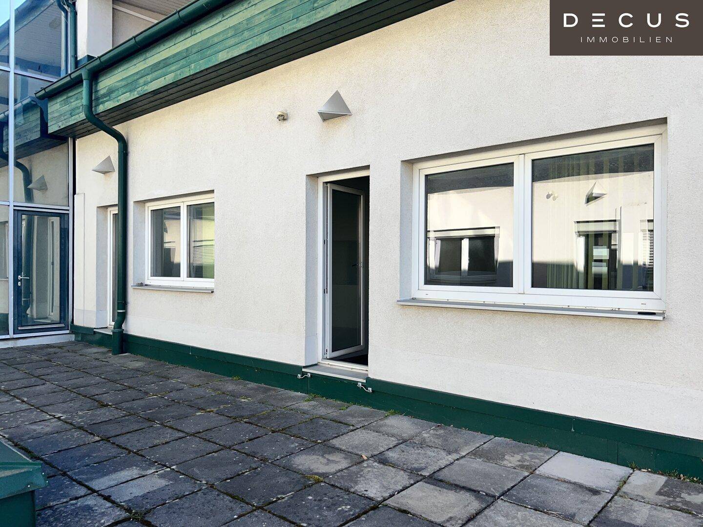 Außenbereich mit gepflasterter Terrasse und weißen Fenstern, die viel Licht in die angrenzenden Räume lassen.