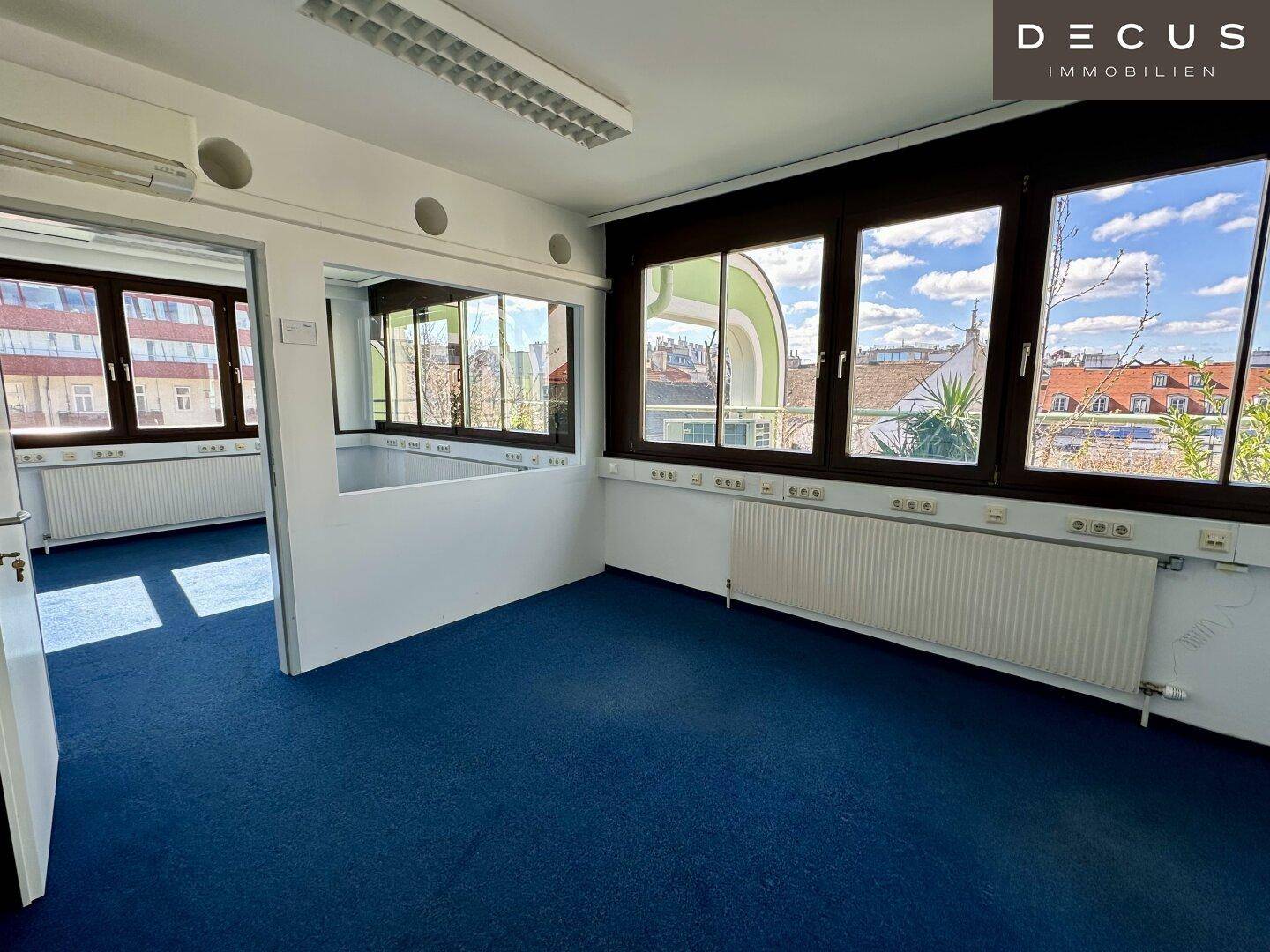 Bürofläche mit großen Fenstern, die einen weiten Blick über die Dächer bieten, und blauem Teppichboden.