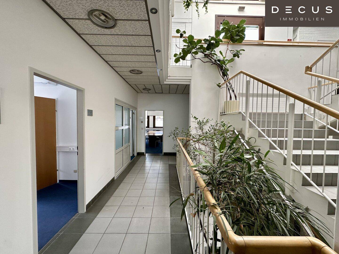Langer Flur mit Fliesenboden, Treppenhaus und Pflanzen, führt zu weiteren Büroräumen.