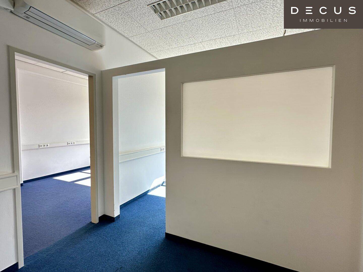 Büroraum mit weißer Wandtafel, blauem Teppichboden und Zugang zu einem weiteren Raum, gut beleuchtet.