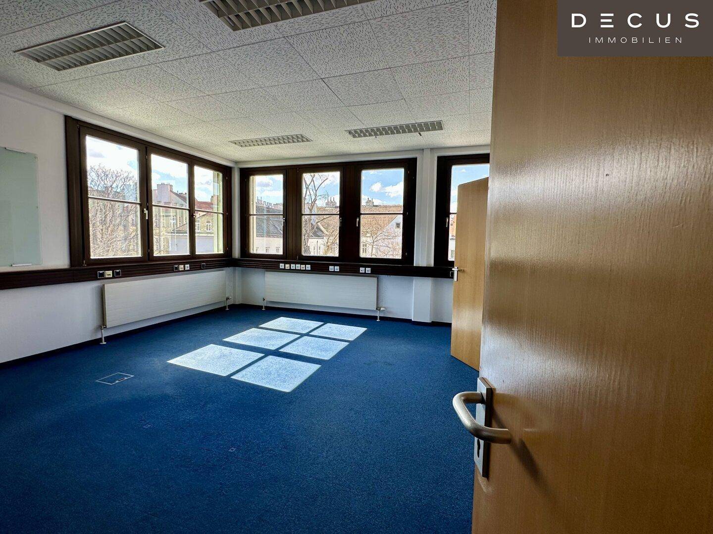 Helles Büro mit blauem Teppichboden, großen Fenstern und Holztür, bietet viel Tageslicht.