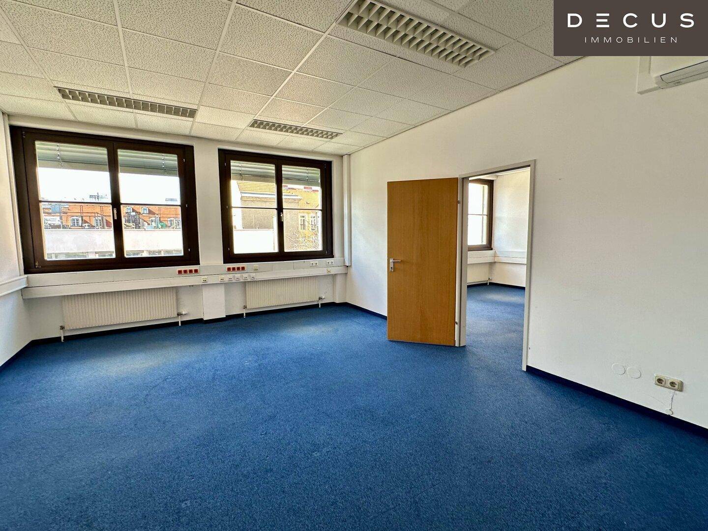 Geräumiges Büro mit blauem Teppichboden und Holztür, bietet eine funktionale Arbeitsumgebung.