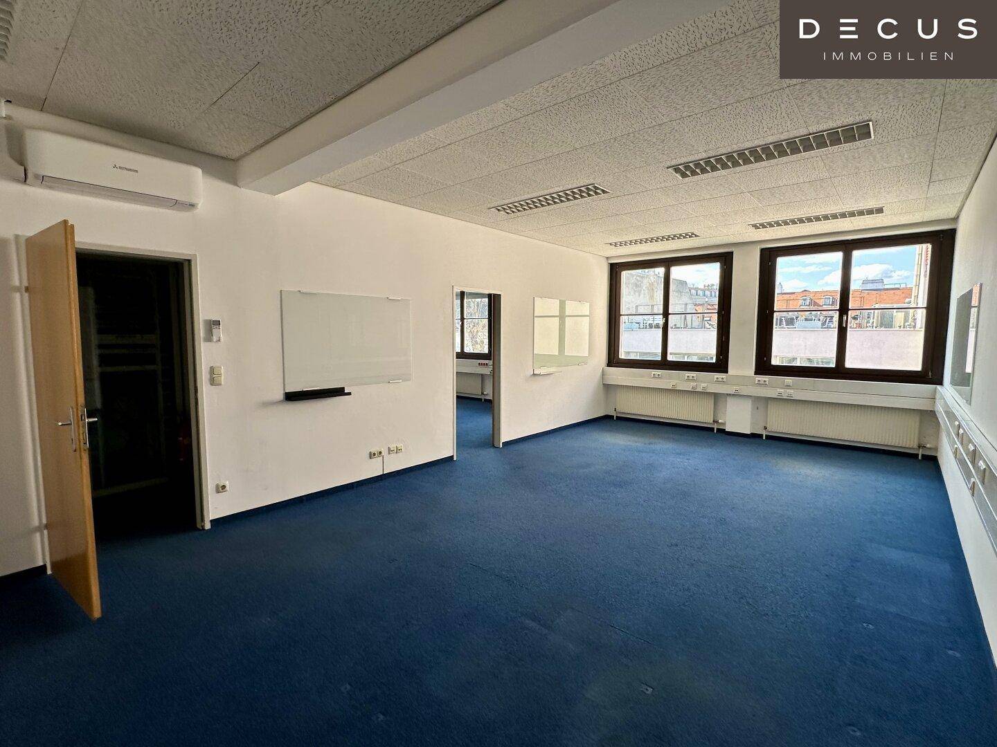 Großes Büro mit blauem Teppichboden, Klimaanlage und vielen Fenstern, ideal für mehrere Arbeitsplätze.