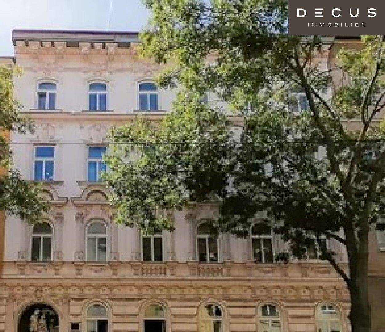 Historische Fassade eines Altbaus mit reich verzierten Fenstern und Stuckelementen, umgeben von Bäumen.