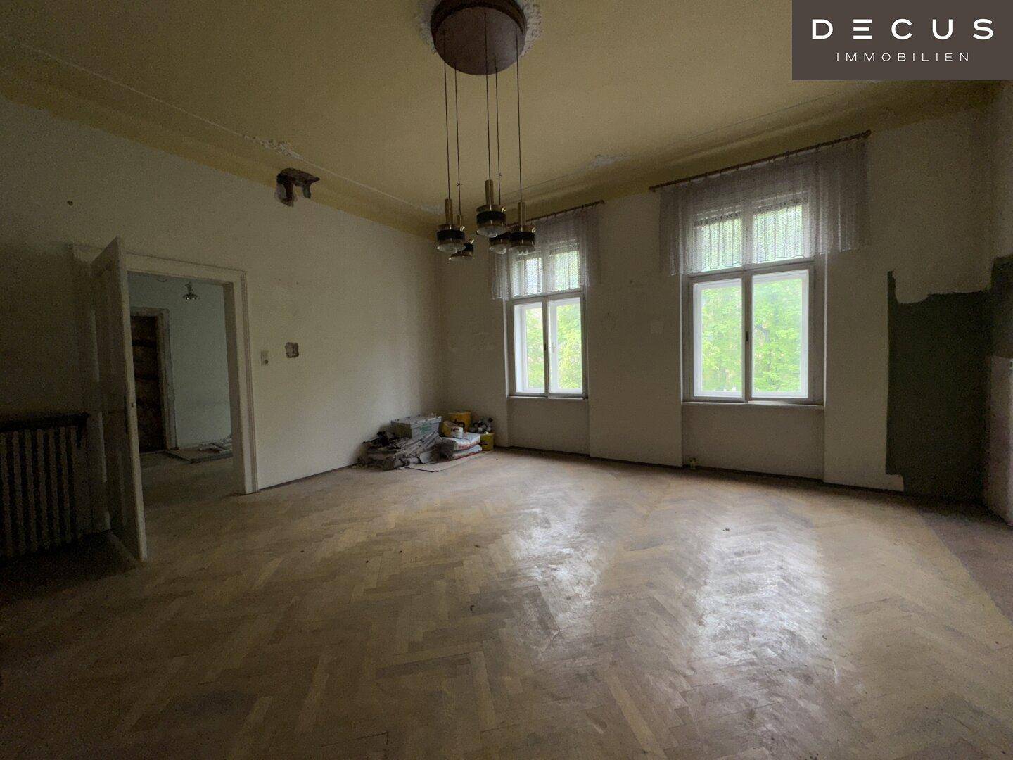 Heller Altbau-Raum mit Fischgrätparkett, hohen Fenstern und Stuckdecke, bereit zur Renovierung.