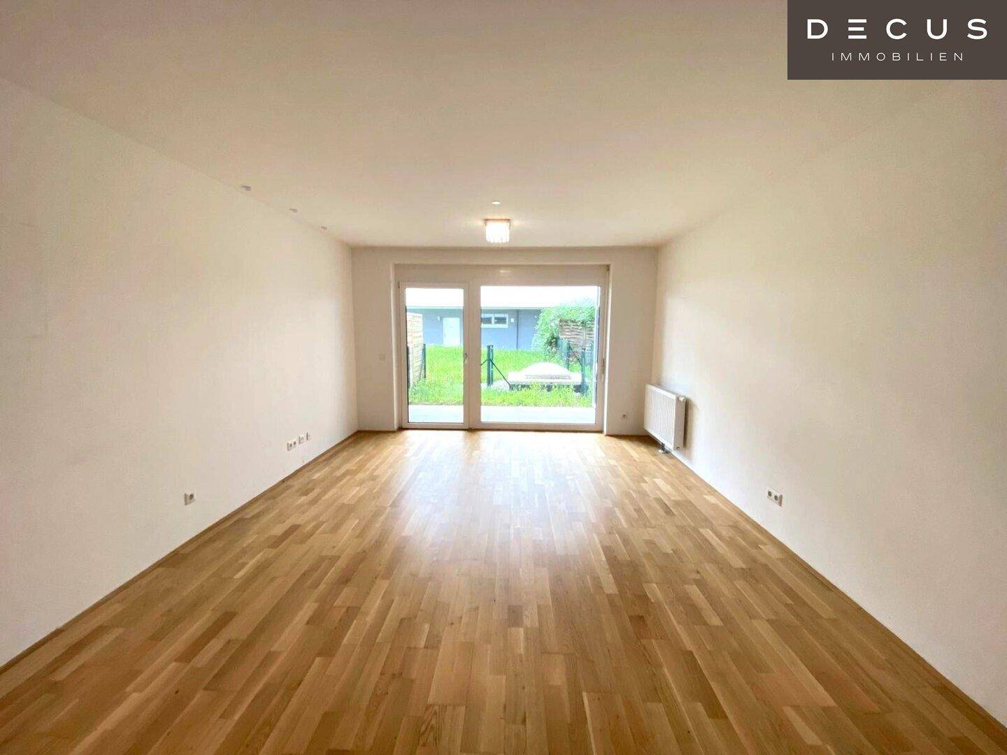 Leerer Raum mit Holzboden und großen Glastüren, die Zugang zu einem Gartenbereich bieten.