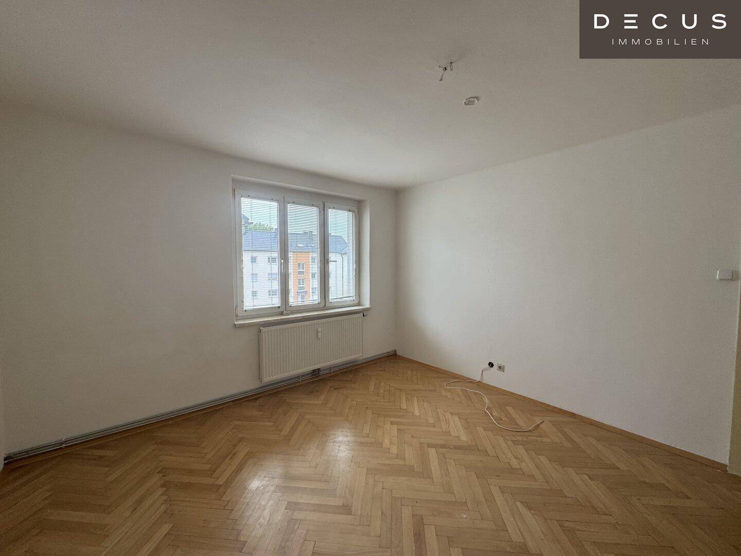 Geräumiges Zimmer mit Fischgrätparkettboden und großem Fenster, bereit zur Renovierung.