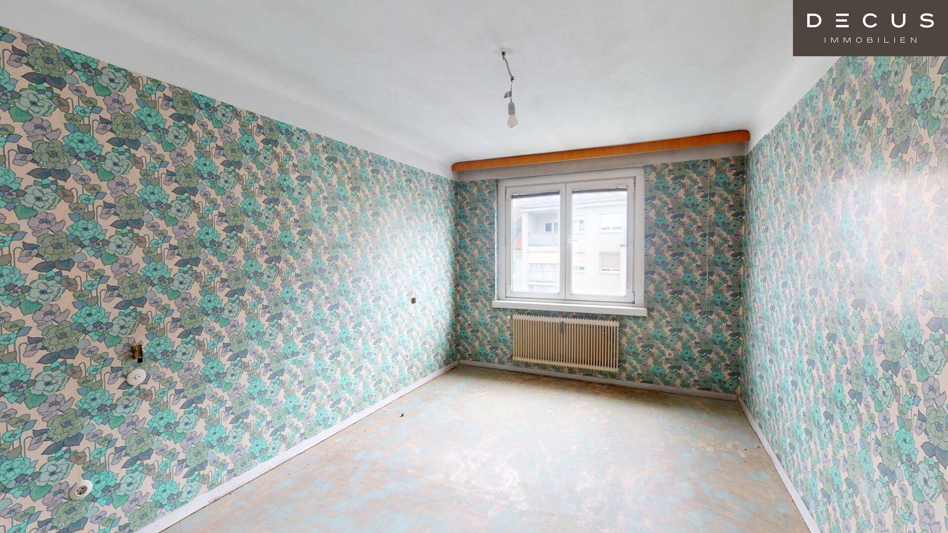 Schlafzimmer mit auffälliger floraler Tapete und Heizkörper unter dem Fenster, benötigt Renovierung.