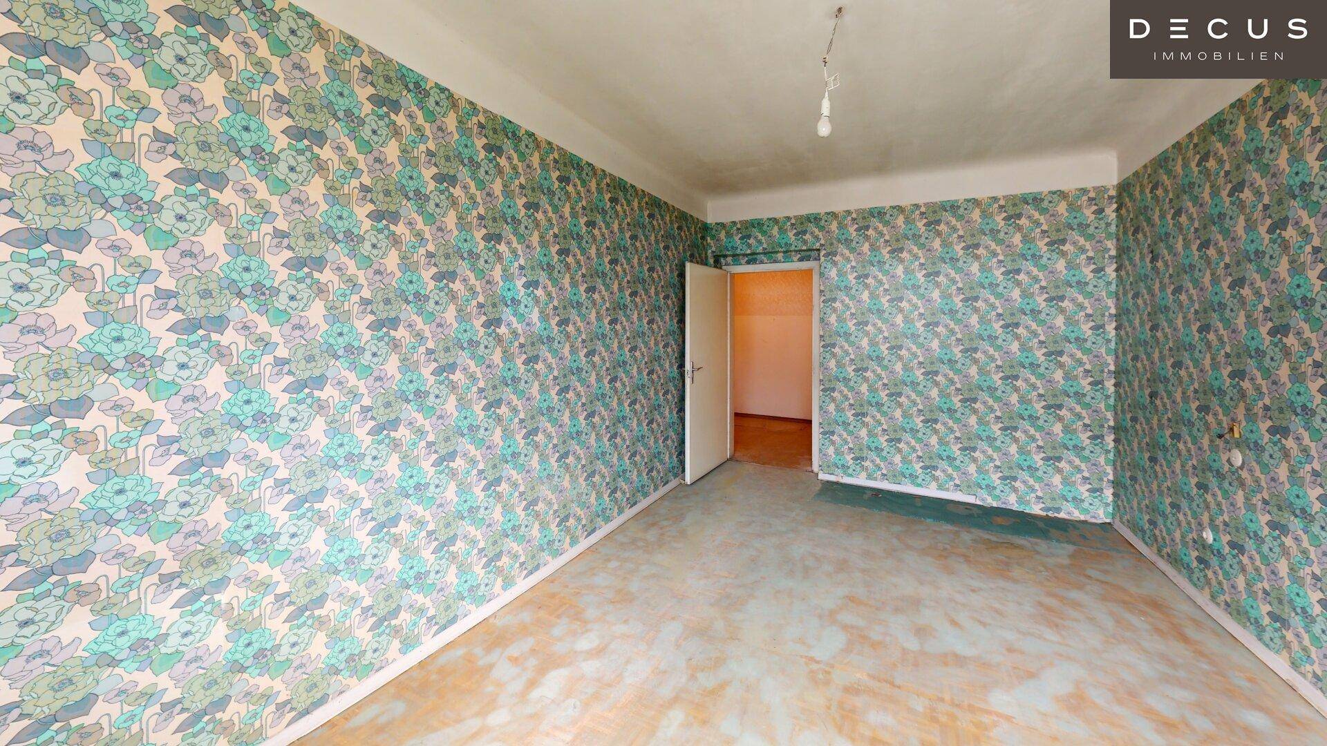 Geräumiges Zimmer mit auffälliger floraler Tapete und Tür, wartet auf eine Renovierung.