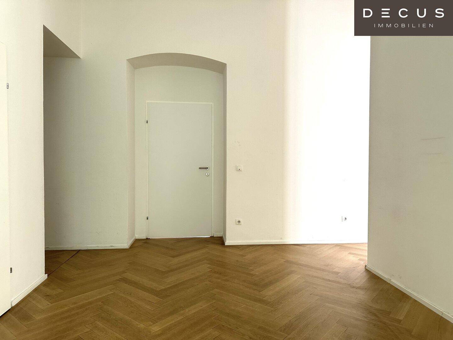 Geräumiger Büroraum mit Fischgrätparkett und einer schlichten weißen Tür in einer Nische, sehr hell.