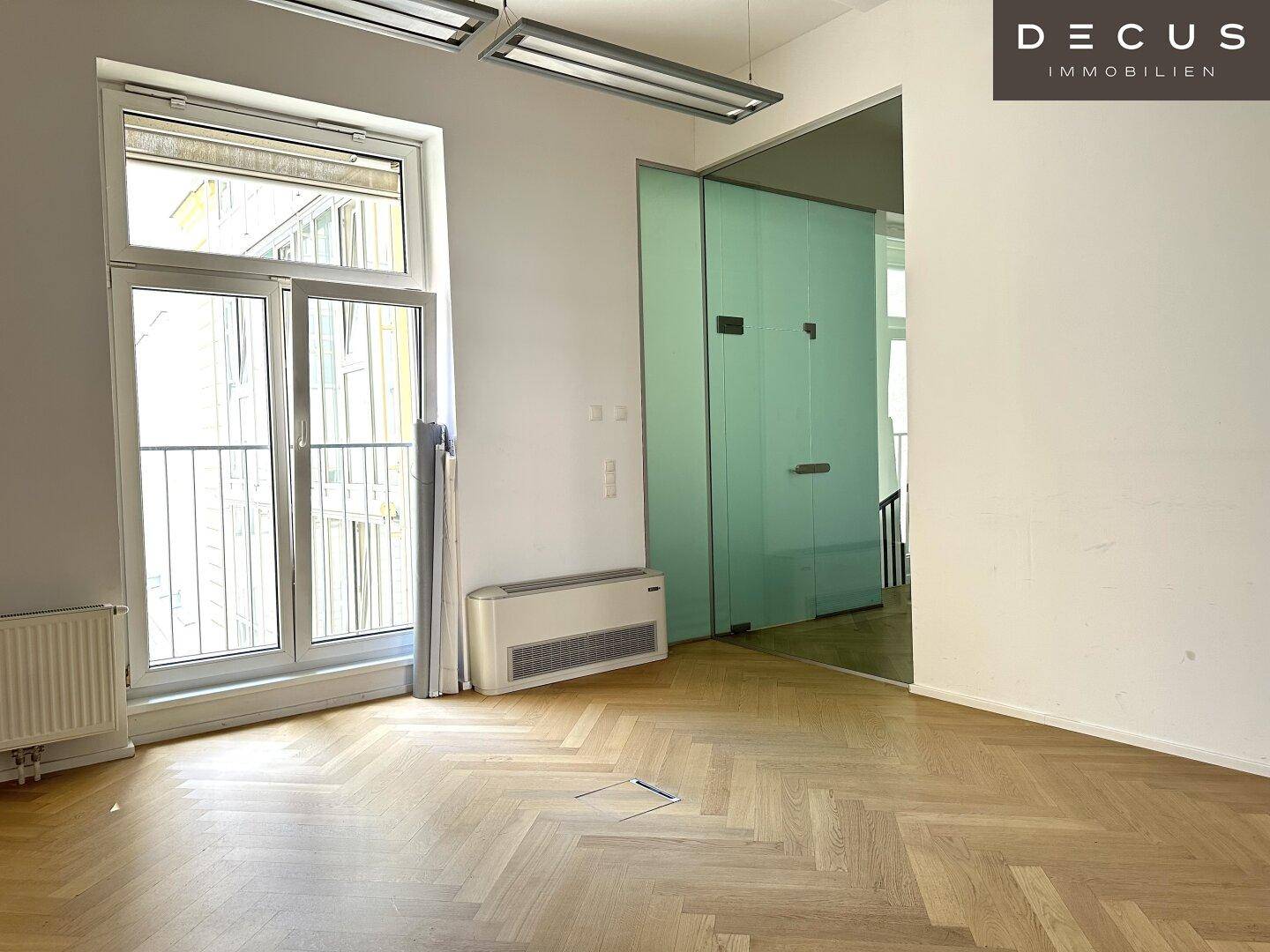 Heller Büroraum mit Fischgrätparkett, großen Fenstern und Zugang zu einem kleinen Balkon, viel Tageslicht.