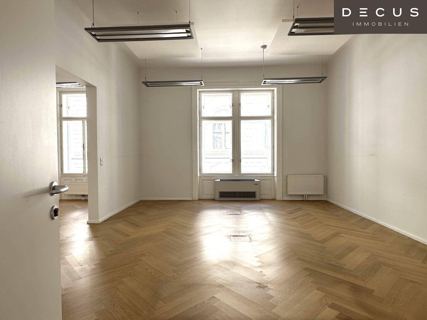 Geräumiger Büroraum mit Fischgrätparkett, großen Fenstern und modernen Deckenleuchten, sehr hell und offen.