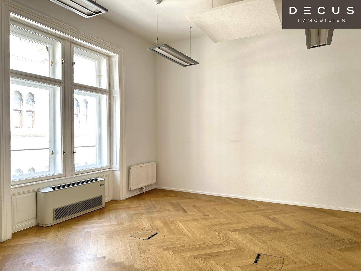 Heller Büroraum mit Fischgrätparkett, großen Fenstern und Blick auf die Straße, viel Tageslicht.