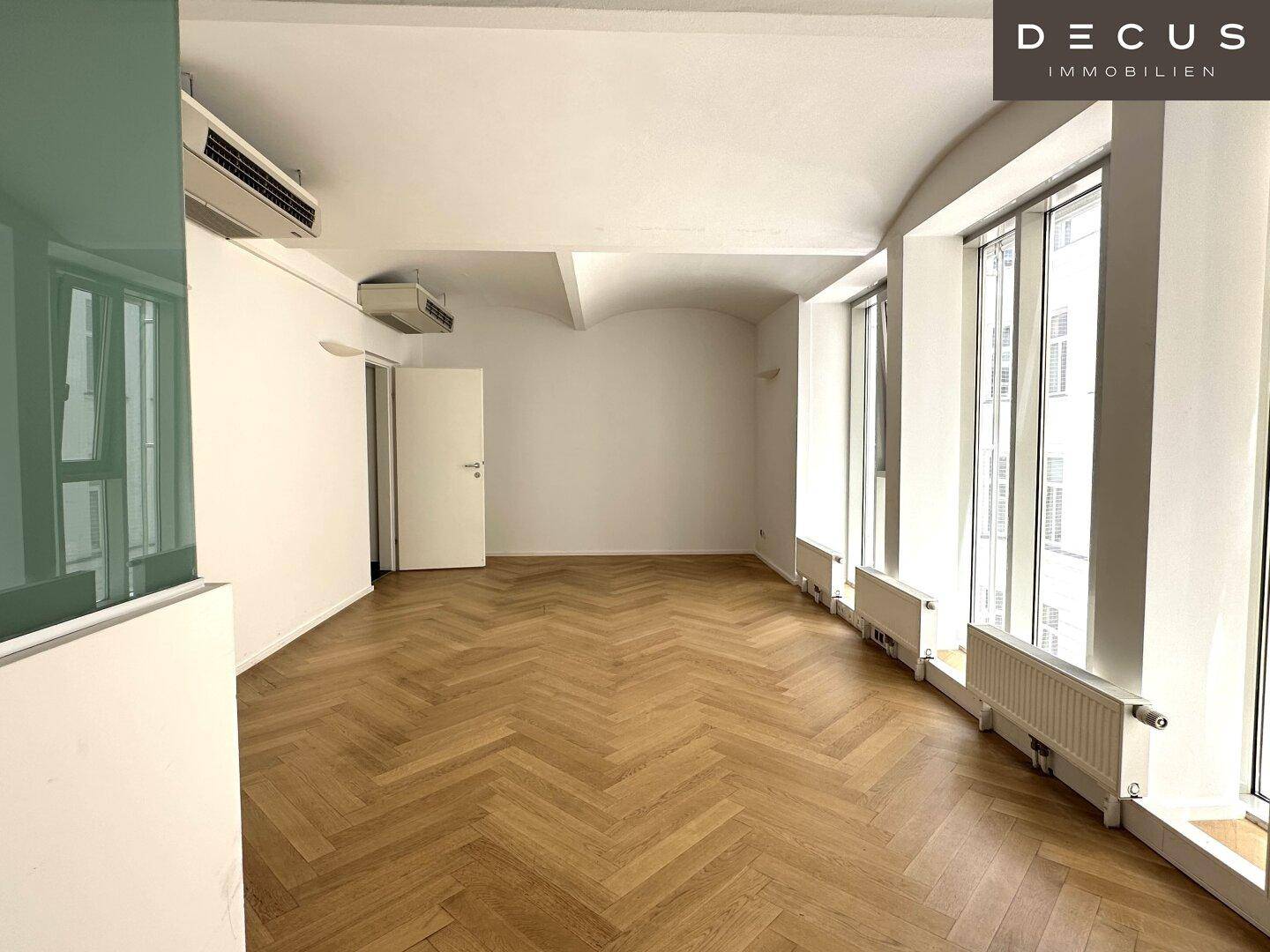 Heller Büroraum mit Fischgrätparkett, hohen Decken und vielen Fenstern, bietet eine offene Arbeitsatmosphäre.