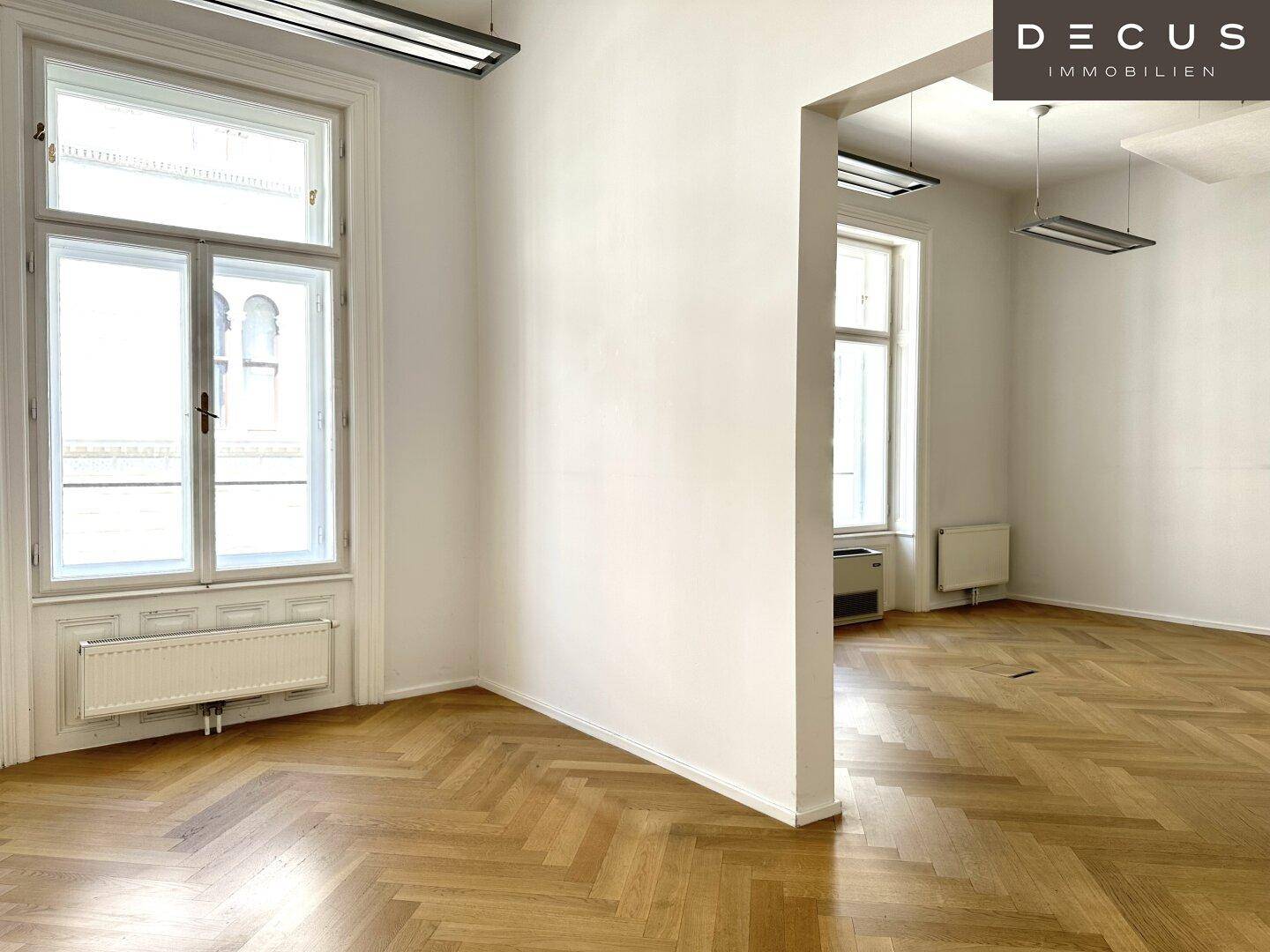 Heller Büroraum mit Fischgrätparkett, großen Fenstern und einem Heizkörper unter dem Fenster, gemütlich und funktional.