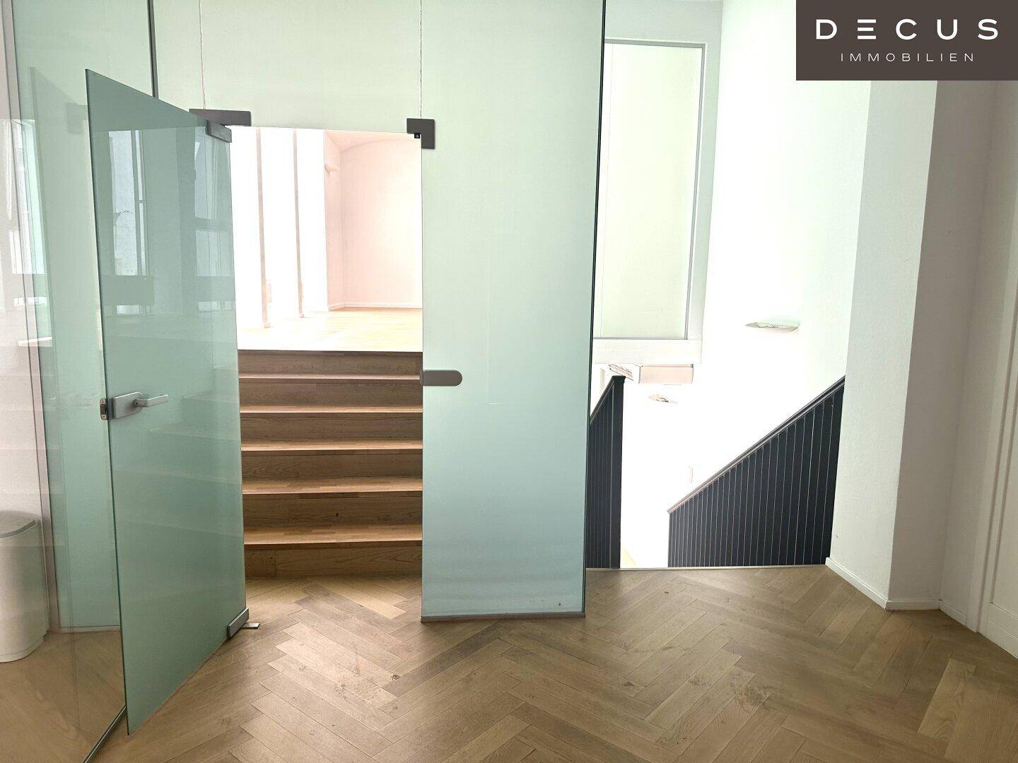Heller Flurbereich mit Glaswänden, Holztreppe und Fischgrätparkett, der verschiedene Ebenen verbindet.