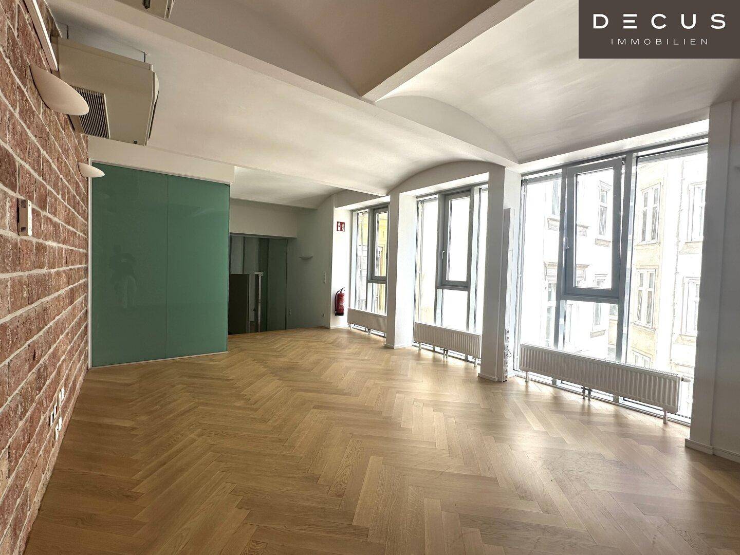 Großzügiger Bürobereich mit Fischgrätparkett, hohen Decken und großen Fenstern, die viel Licht hereinlassen.