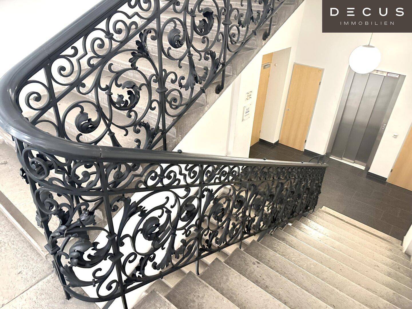 Elegantes Treppenhaus mit schmiedeeisernem Geländer und Zugang zu einem modernen Aufzug in einem Altbau.