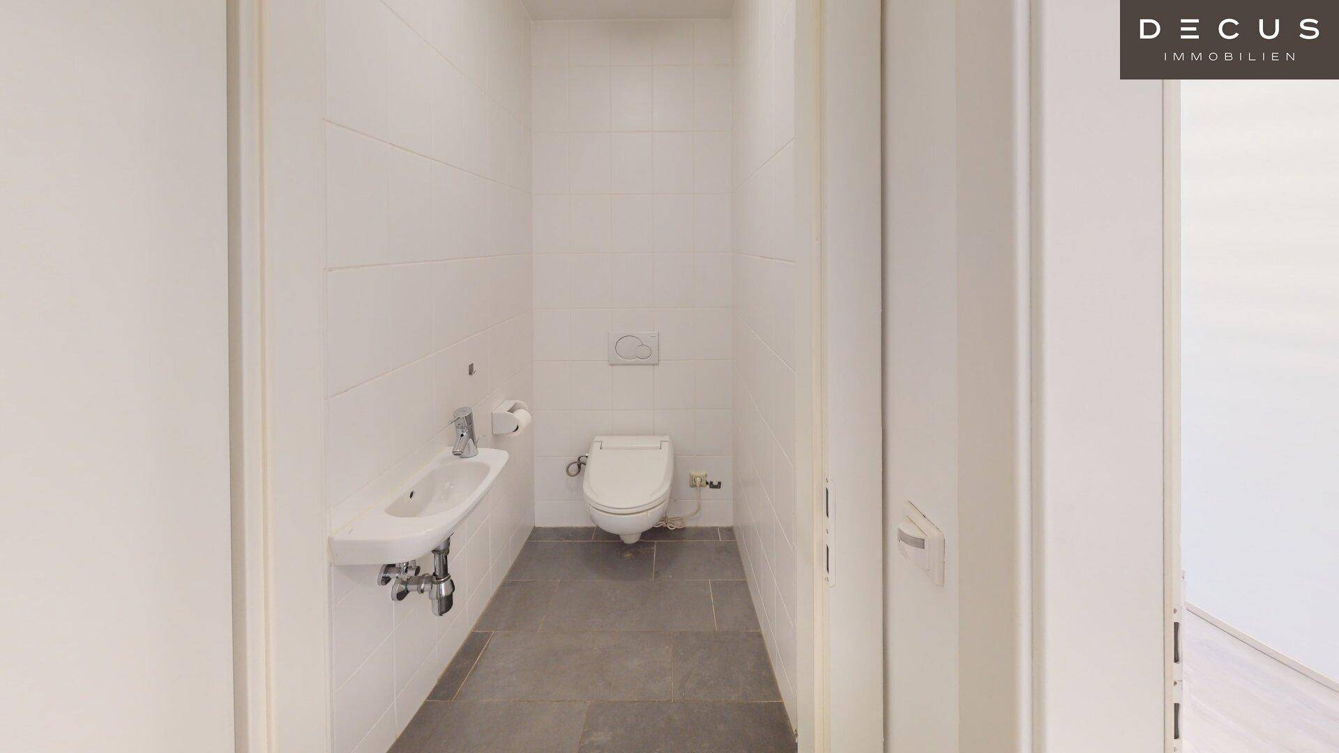 Helles Gäste-WC mit Wand-WC, kleinem Waschbecken und modernen, hellen Fliesen.