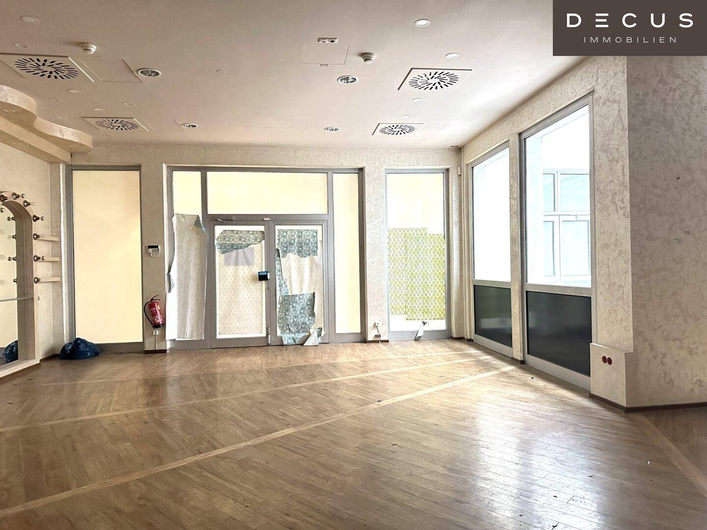 Geräumiger, leerer Gewerberaum mit großen Fenstern, Holzboden und Lüftungsanlagen an der Decke.