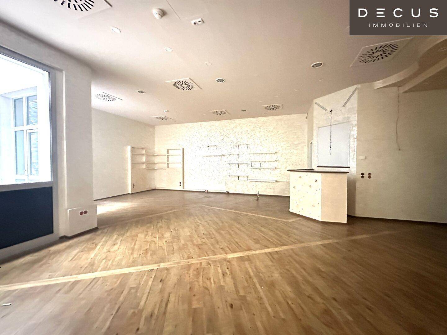 Weitläufiger Gewerberaum mit Holzboden, vorbereiteten Regalsystemen und einer kleinen Theke.